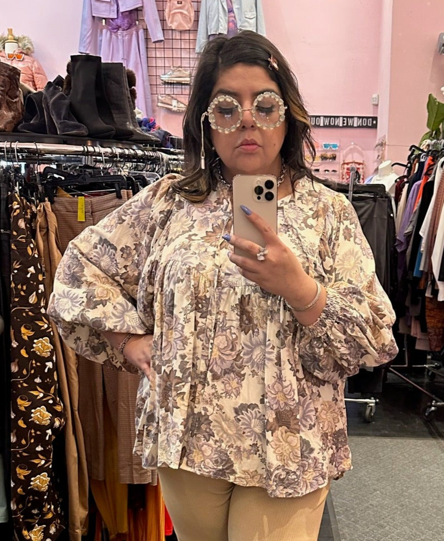 Close up view of a size XXL The Lullaby Club neutral tone floral blouse with fabric buttons, puff sleeves, and a baby doll shape styled with khaki trousers and floral glasses on a size 14/16 model.