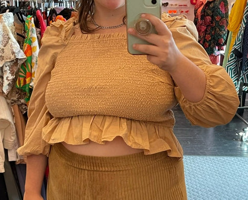 Close up front view of a size XL Amazon The Drop neutral tone tan smocked long puff sleeve crop top with ruffle hem styled with dark yellow corduroy pants on a size 14/16 model.