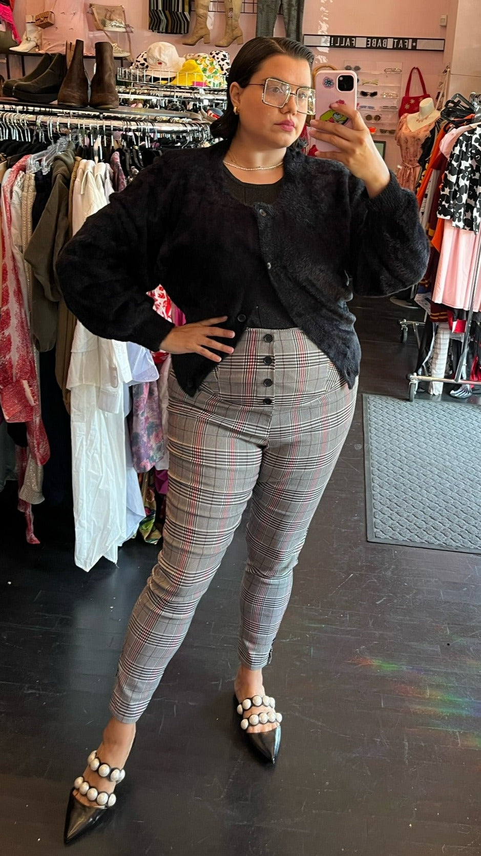 Full-body front view of a pair of size 2X Unique Vintage black, white, and red plaid legging-style high-waisted pants with button details styled with a black fuzzy cardigan and pearl-studded slides on a size 14/16 model.