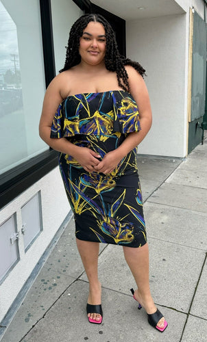 Full-body front view of a size 3X Rue 107 black strapless ruffle bust bodycon column dress with all-over yellow, teal, and royal blue paint splatter pattern styled with black heels on a size 18/20 model. The photo is taken outside in natural lighting.