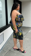 Load image into Gallery viewer, Full-body side view of a size 3X Rue 107 black strapless ruffle bust bodycon column dress with all-over yellow, teal, and royal blue paint splatter pattern styled with black heels on a size 18/20 model. The photo is taken outside in natural lighting.
