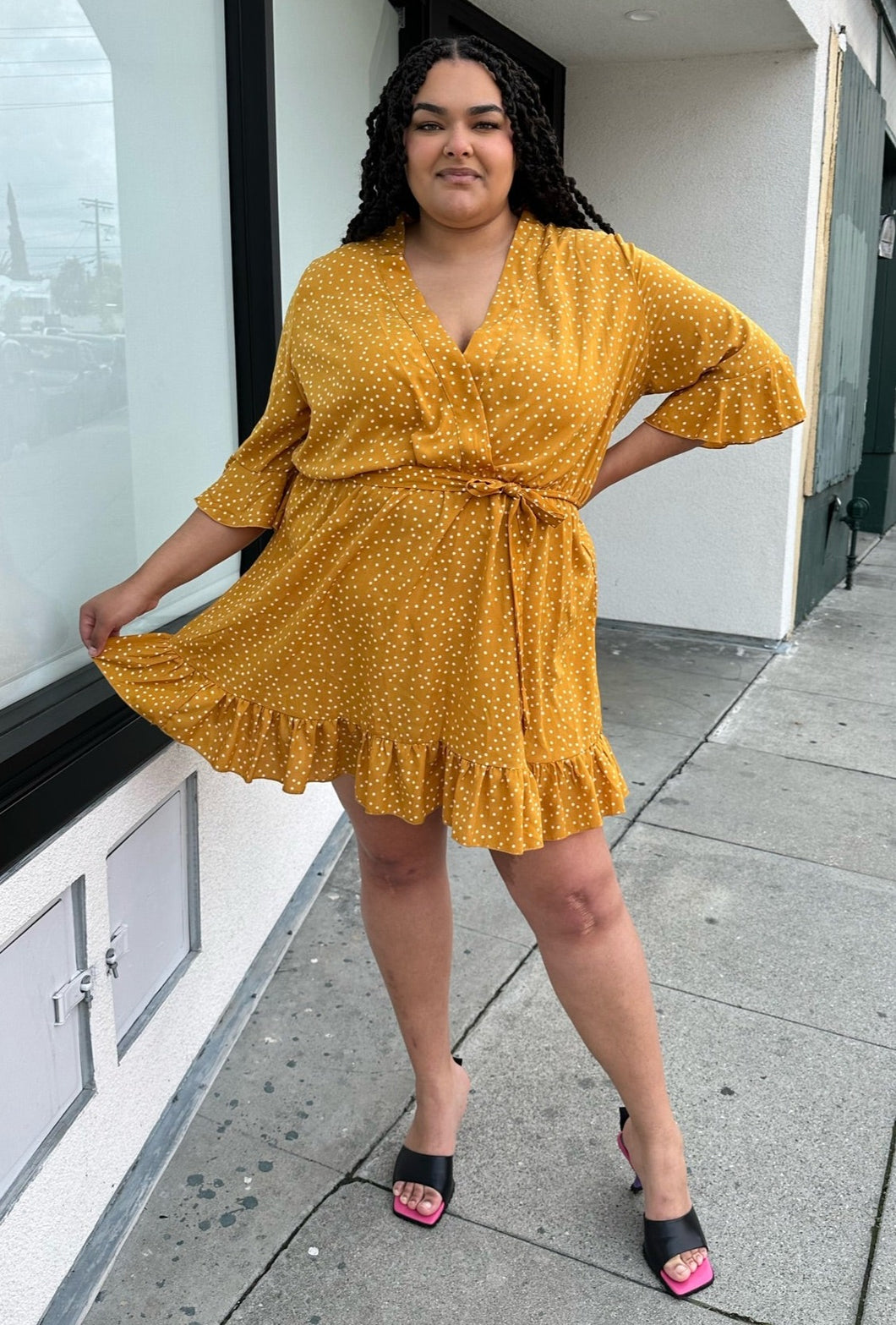 Full-body front view showing off the skirt hem of a size 20 Boohoo mustard yellow faux wrap mini dress with white polka dots, a ruffle hem, and a subtle bell sleeve styled with black heels on a size 18/20 model.