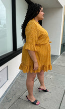 Load image into Gallery viewer, Full-body side view of a size 20 Boohoo mustard yellow faux wrap mini dress with white polka dots, a ruffle hem, and a subtle bell sleeve styled with black heels on a size 18/20 model.
