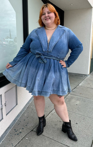 Full-body front view of a size 3 Fashion to Figure x Patrick Starrr lightwash to mediumwash ombré denim belted mini dress with collar and hook-and-eye bust detail styled with black boots on a size 22/24 model.