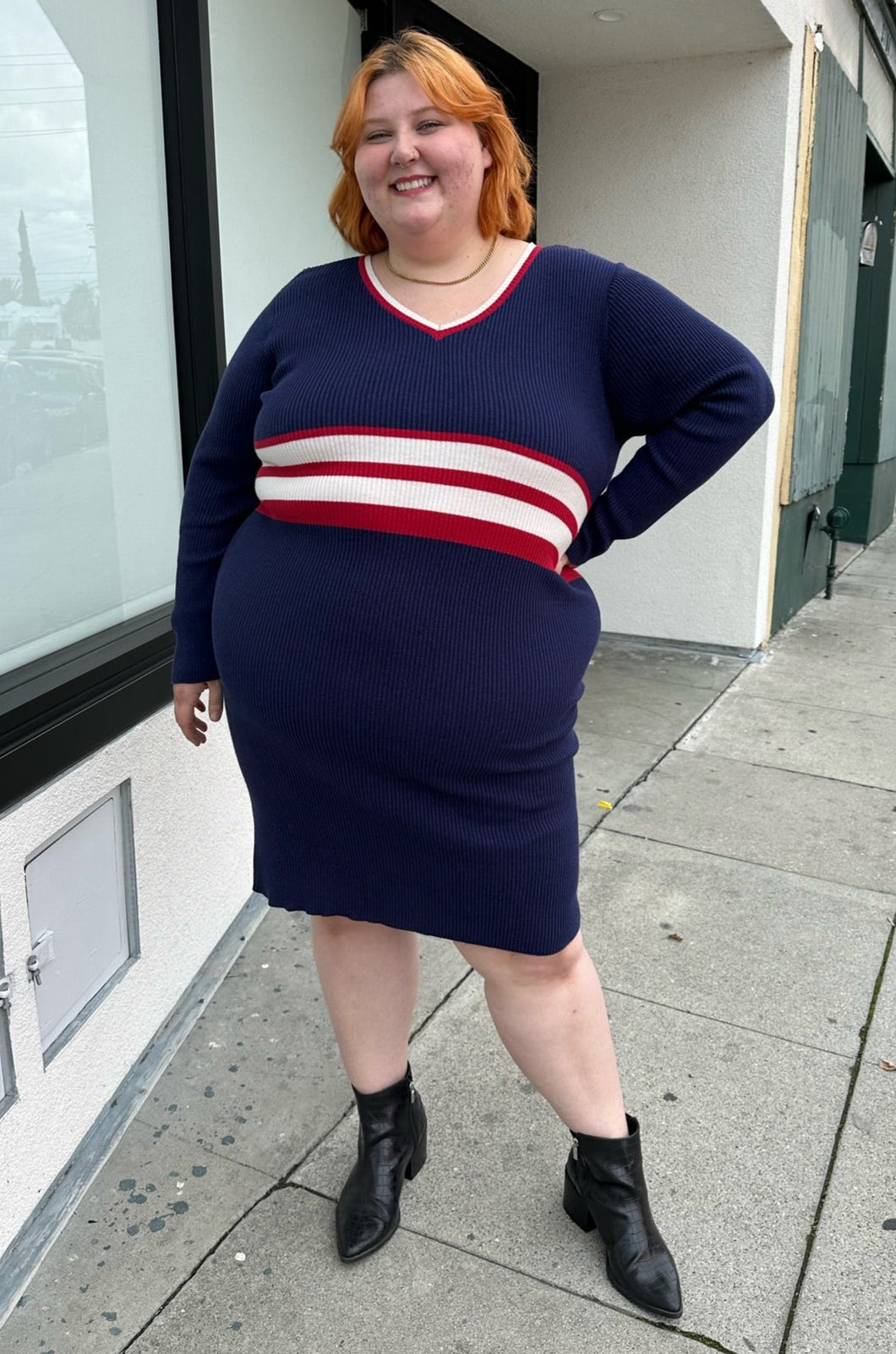 Full-body front view of a size 22/24 Eloquii x Katie Sturino navy blue ribbed knit sweater dress with red and white varisty stripes and a red and white v-neck stripe styled with black boots on a size 22/24 model. The photo is taken outside in natural lighting.