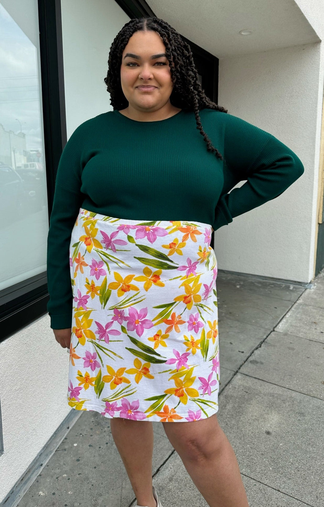 Front view of a size 2X Unicorn Tears vintage white midi skirt with pink, yellow, and green floral pattern styled with a deep emerald long sleeve top on a size 18/20. The photo is taken outside with natural lighting.
