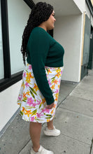 Load image into Gallery viewer, Side view of a size 2X Unicorn Tears vintage white midi skirt with pink, yellow, and green floral pattern styled with a deep emerald long sleeve top on a size 18/20. The photo is taken outside with natural lighting.
