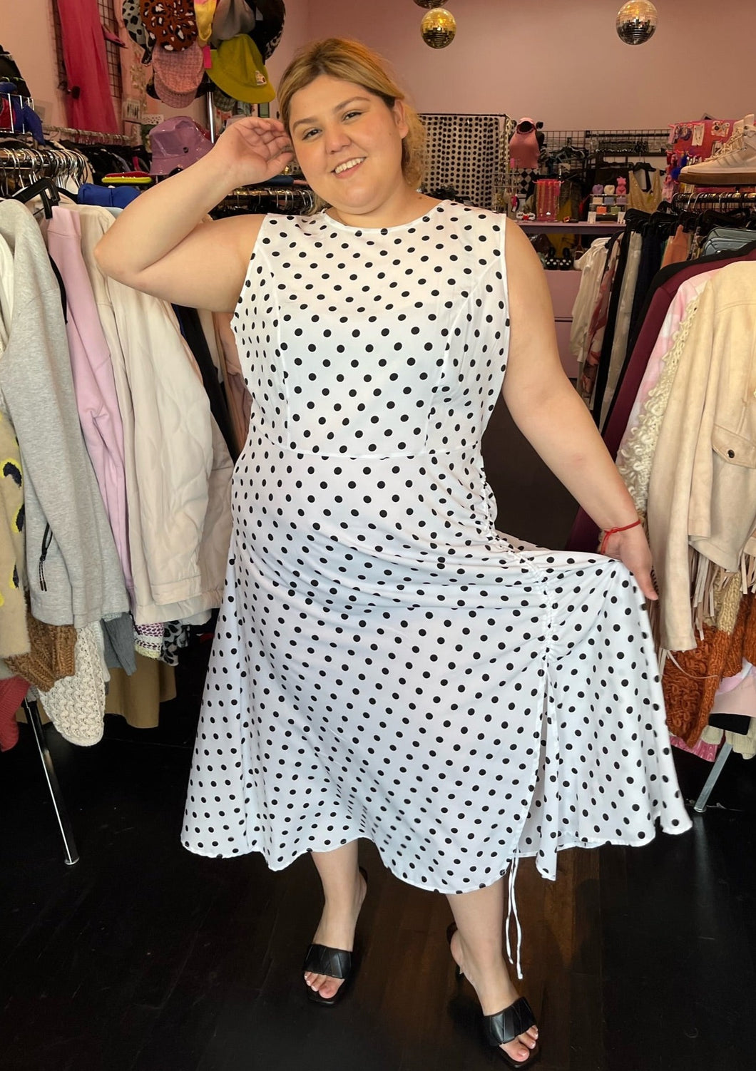 Full-body front view showing off the length of a size 18 ASOS white sleeveless maxi dress with black polka dots and a ruched slit detail at the hem styled with black heels on a size 14/16 model. The photo is taken inside in front of a window, combining natural lighting and overhead lighting.