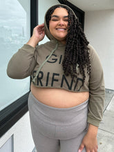 Load image into Gallery viewer, Front view of a size 2X Onuolayichu olive green cropped hoodie that reads, &quot;GIRLFRIEND&quot; styled with the hood up and gray leggings on a size 18/20 model.
