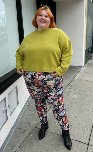 Full-body front view of a pair of size 3X Miss Look muted multicolor floral leggings styled with a chartreuse crewneck and black boots on a size 22/24 model.