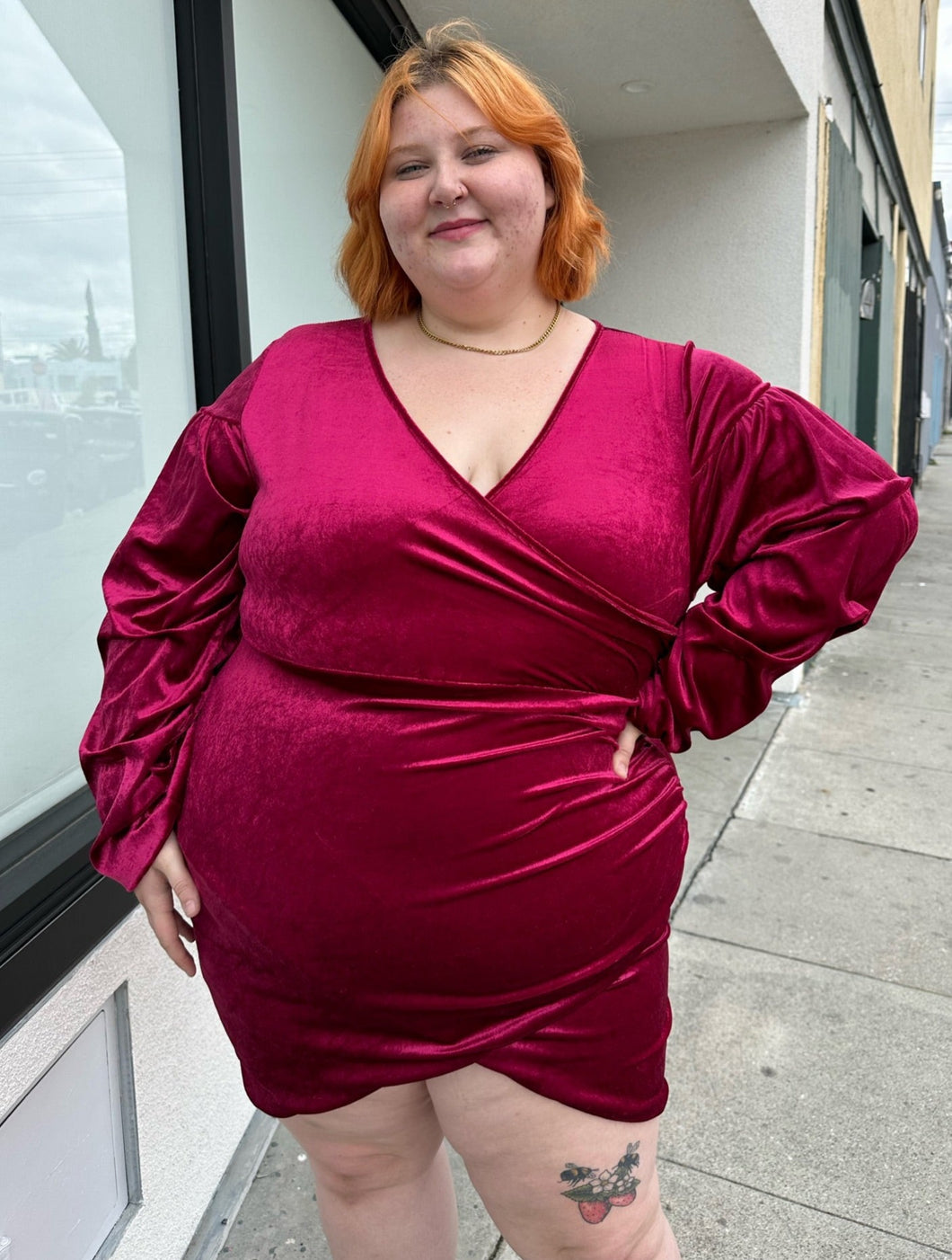 Front view of a size 22 Bloomchic deep red velvet mini dress with draped details and long, puff sleeves on a size 22/24 model. This photo is taken outside in natural lighting.