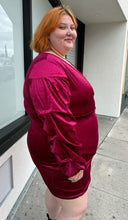 Load image into Gallery viewer, Side view of a size 22 Bloomchic deep red velvet mini dress with draped details and long, puff sleeves on a size 22/24 model. This photo is taken outside in natural lighting.
