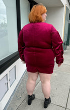 Load image into Gallery viewer, Full-body back view of a size 22 Bloomchic deep red velvet mini dress with draped details and long, puff sleeves styled with black boots on a size 22/24 model. This photo is taken outside in natural lighting.
