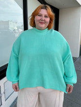 Load image into Gallery viewer, Additional front view of a size 18 ASOS cyan teal thick knit mockneck oversized sweater styled over cream pants on a size 22/24 model.
