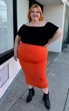 Load image into Gallery viewer, Full-body front view of a size 3X Fashion Nova bright orange bodycon maxi skirt styled with a black off-shoulder blouse and black pointy boots on a size 22/24 model.

