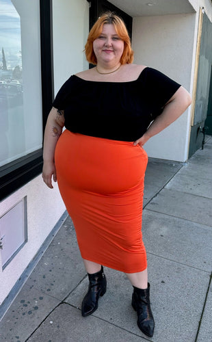 Full-body front view of a size 3X Fashion Nova bright orange bodycon maxi skirt styled with a black off-shoulder blouse and black pointy boots on a size 22/24 model.