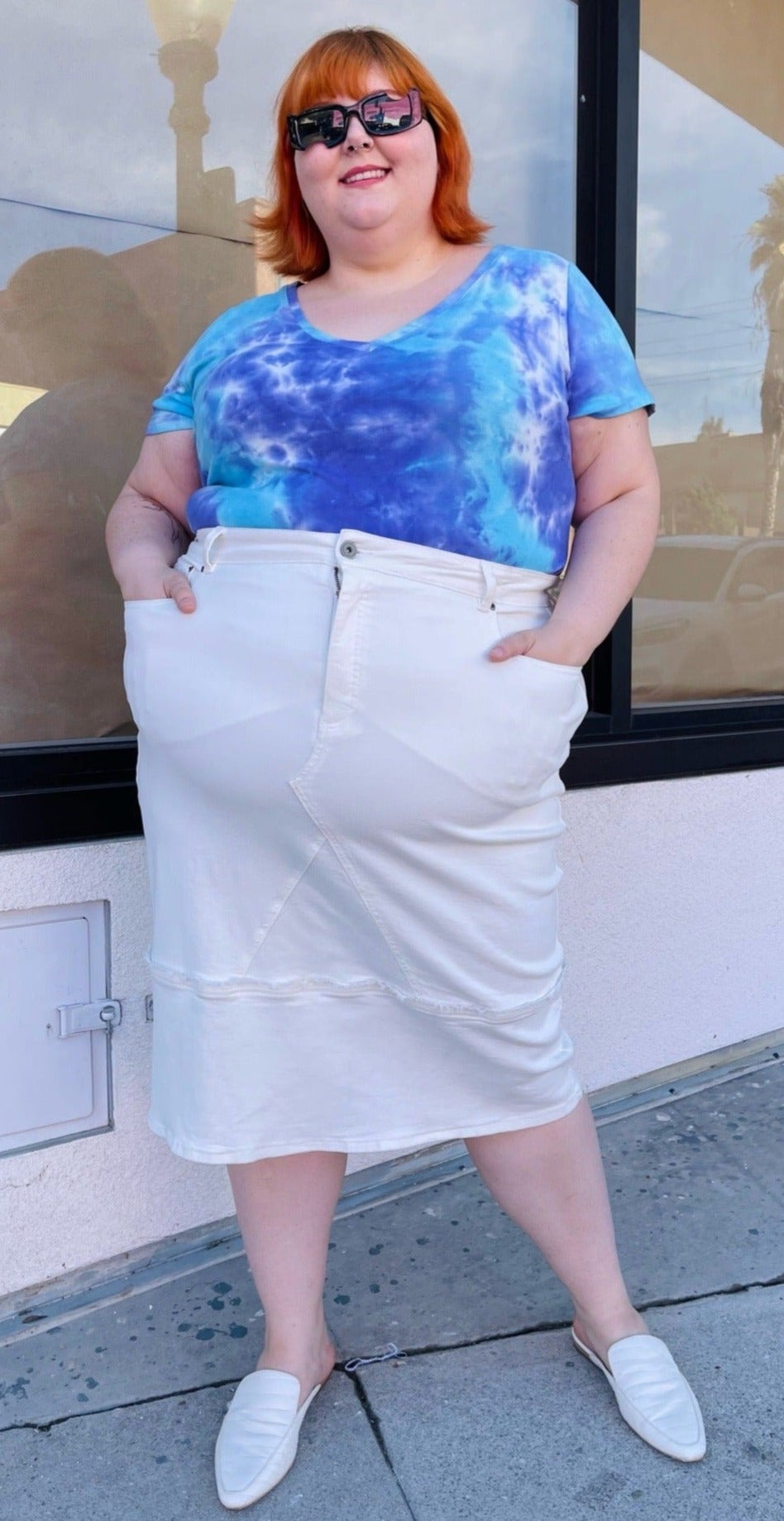 Full-body front view of a size L Universal Standard white deconstructed denim midi skirt styled with a blue and purple tie dye tee and white slides on a size 22/24 model.