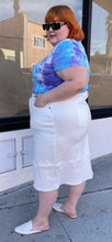Load image into Gallery viewer, Full-body side view of a size L Universal Standard white deconstructed denim midi skirt styled with a blue and purple tie dye tee and white slides on a size 22/24 model.
