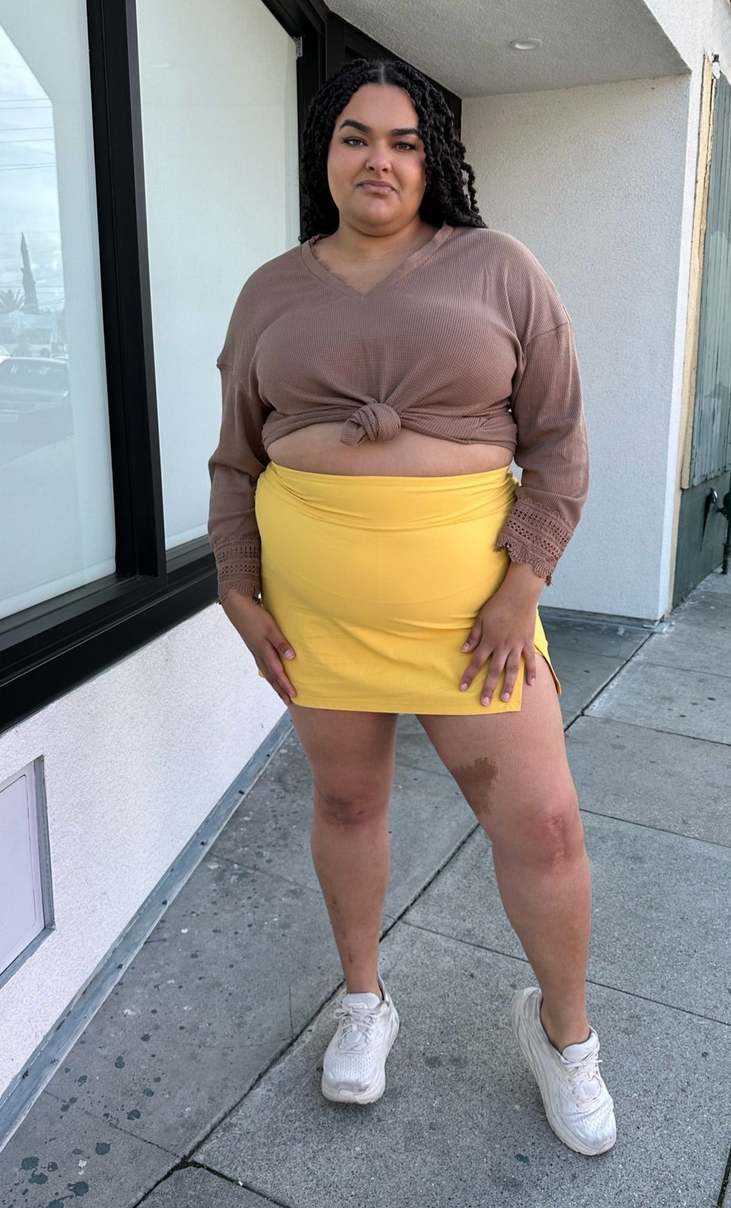 Full-body front view of a size 2X Alder sunshine yellow mini skirt with a small front-side slit detail styled with a brown sweater and white sneakers on a size 18/20 model.