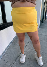 Load image into Gallery viewer, Front view of a size 2X Alder sunshine yellow mini skirt with a small front-side slit detail styled with a brown sweater and white sneakers on a size 18/20 model.
