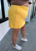Load image into Gallery viewer, Side view of a size 2X Alder sunshine yellow mini skirt with a small front-side slit detail styled with a brown sweater and white sneakers on a size 18/20 model.
