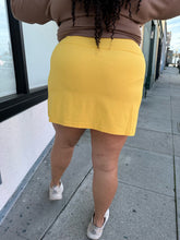 Load image into Gallery viewer, Back view of a size 2X Alder sunshine yellow mini skirt with a small front-side slit detail styled with a brown sweater and white sneakers on a size 18/20 model.
