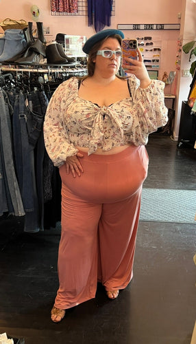 Full-body front view of a pair of size 3X Zelie for She dusty pink stretch high-waisted pants styled with a white floral romantic blouse, a blue beret, and blue sunnies on a size 24 model.