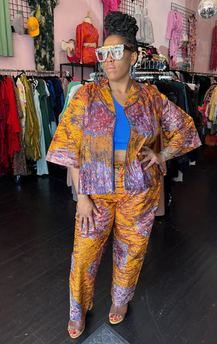 Full-body front view of a size 16 Le Victoria for RAYO orange, purple, blue, and maroon two-piece cotton button-up shirt and trouser set styled open over a cobalt blue crop top with heels and bejeweled glasses. The model stands in front of natural light and under overhead lights indoors.