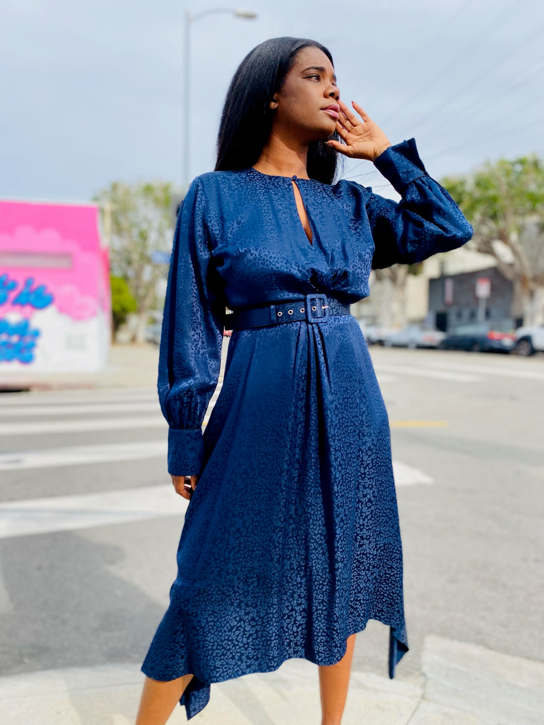 Photo of a size 10/12 model standing outside in front of brightly colored buildings wearing a size 14 Jonathan Simkhai belted navy blue leopard print burnout midi dress with a handkerchief hemline, a keyhole bust detail, and a subtle balloon sleeve.