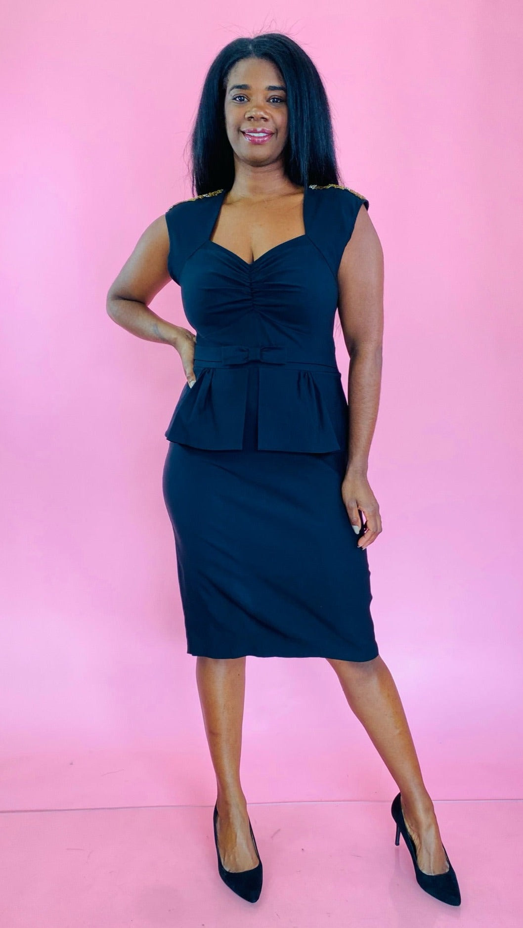 Full-body front view of a size 18 Stop Staring black peplum midi dress with square-sweetheart neckline, ruching at the bust, and a bow detail at the waist styled with black pumps on a size 10/12 model.