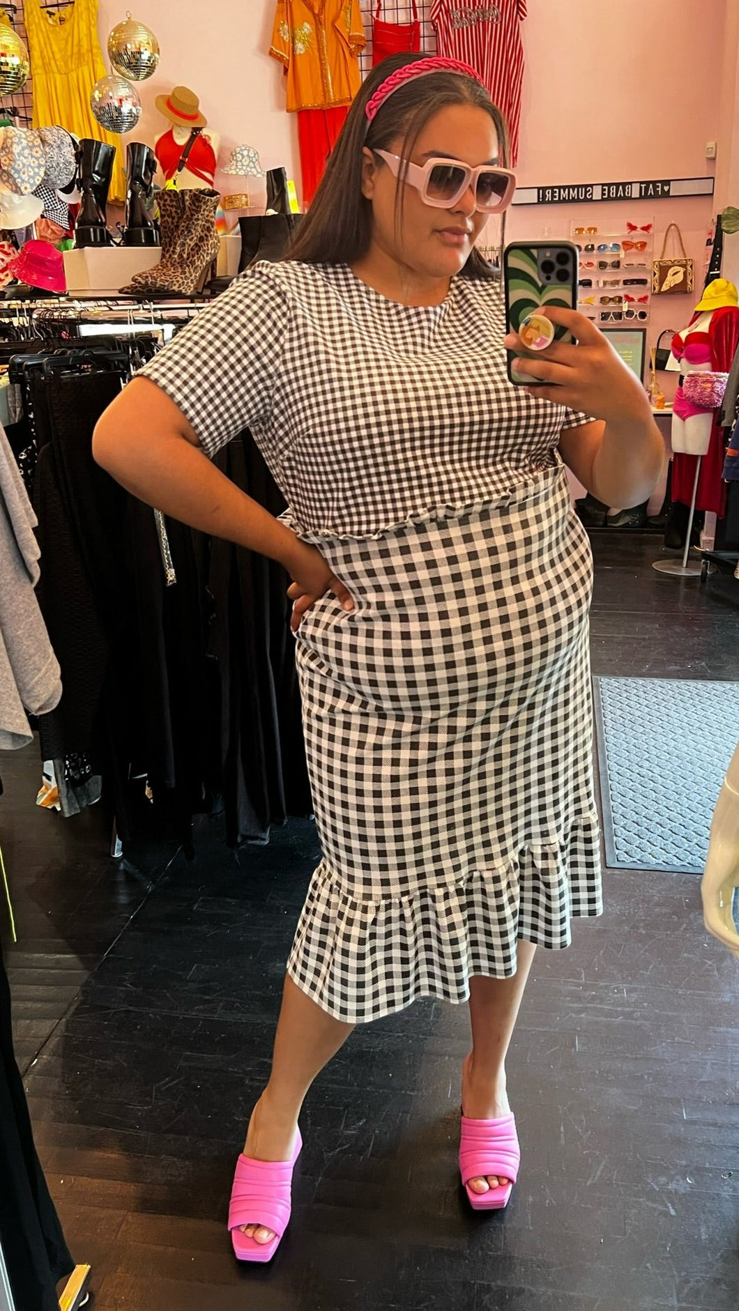 Full-body front view of a size 18 ASOS black and white mixed pattern gingham sheath dress with a mini ruffle at the waist and a larger ruffle at the hem styled with a pink headband and pink mules on a size 18/20 model.