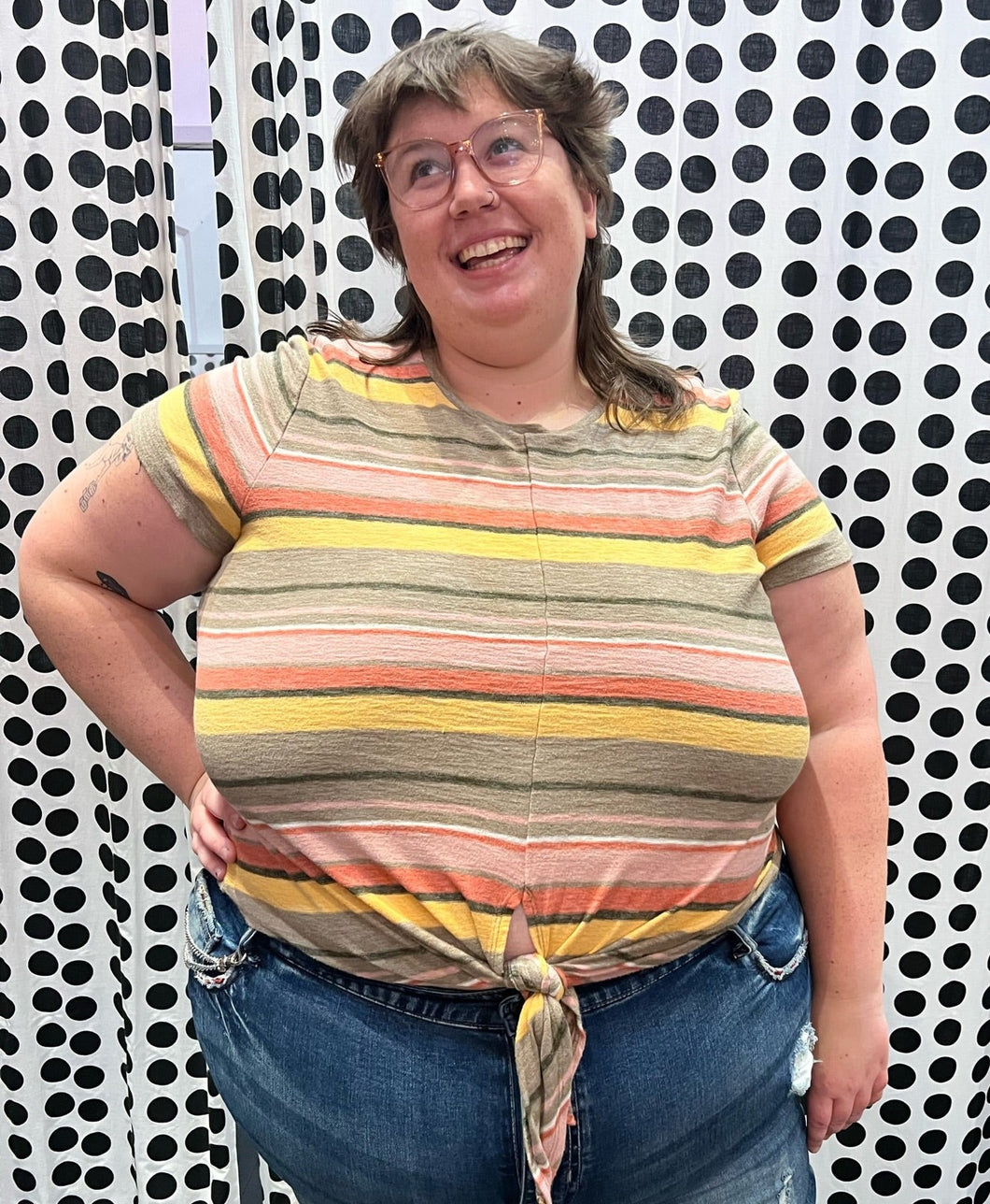 Front view of a size 3X Texture & Thread (Madewell) orange, pink, brown, and cream horiztonal striped tee shirt with a tie detail at the bottom styled with medium-wash denim on a size 24 model.