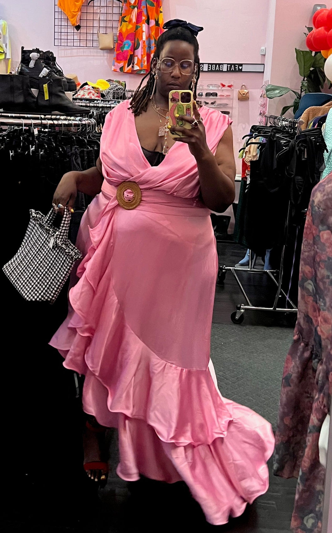 Full-body front view of a size 4 Fashion to Figure x Garner Style collab bubblegum pink maxi wrap dress with big ruffle tiers and a wicker-style buckle belt styled with a gingham print handbag on a size 24/26 model.