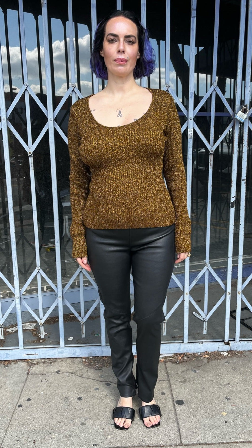 Full-body front view of a size XL (fits up to size 24) GANNI deep gold metallic ribbed knit sweater styled with leather pants and black slides on a size 10 model. The photo is taken outside in natural lighting.