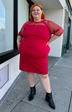 Load image into Gallery viewer, Additional full-body side view of a size 24 Lane Bryant red sheath midi dress with a faux-halter style neckline and long sheer mesh swiss dot sleeves styled with gold jewelry and black boots on a size 22/24 model.
