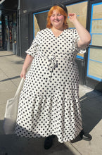 Load image into Gallery viewer, Full-body front view of a size 4X Rebdolls white and black polka dot ruffle-tiered maxi dress with ruffle sleeves and tie belt styled with leather boots and a leather hat on a size 22/24 model. Photo is taken outside in the sun.
