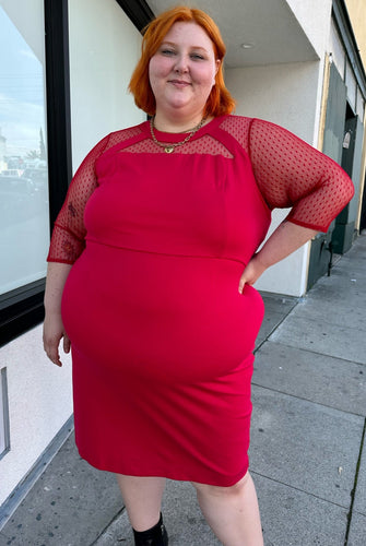 Front view of a size 24 Lane Bryant red sheath midi dress with a faux-halter style neckline and long sheer mesh swiss dot sleeves styled with gold jewelry and black boots on a size 22/24 model.