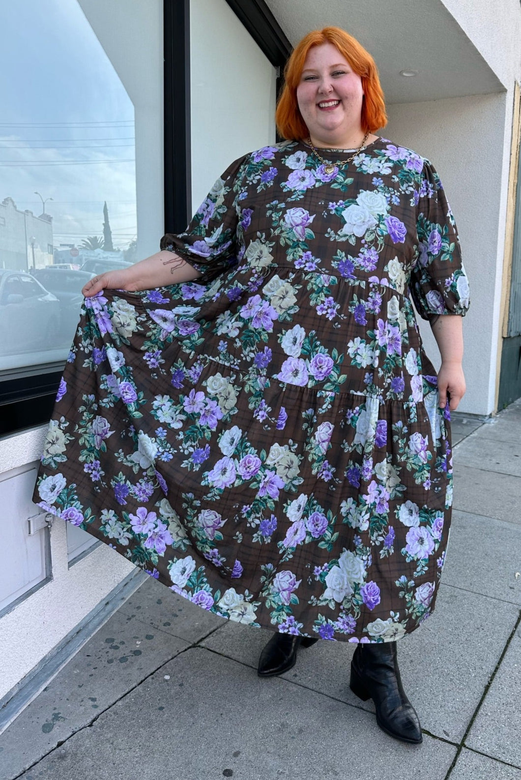 Full-body front view of a size 26 ASOS dark olive green, vibrant green, and lavender mixed print floral and plaid tiered maxi dress with puff sleeves styled with black boots on a size 22/24 model. The photo is taken outside in natural lighting.
