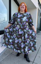 Load image into Gallery viewer, Full-body front view showing off the flounce of the skirt of a size 26 ASOS dark olive green, vibrant green, and lavender mixed print floral and plaid tiered maxi dress with puff sleeves styled with black boots on a size 22/24 model. The photo is taken outside in natural lighting.
