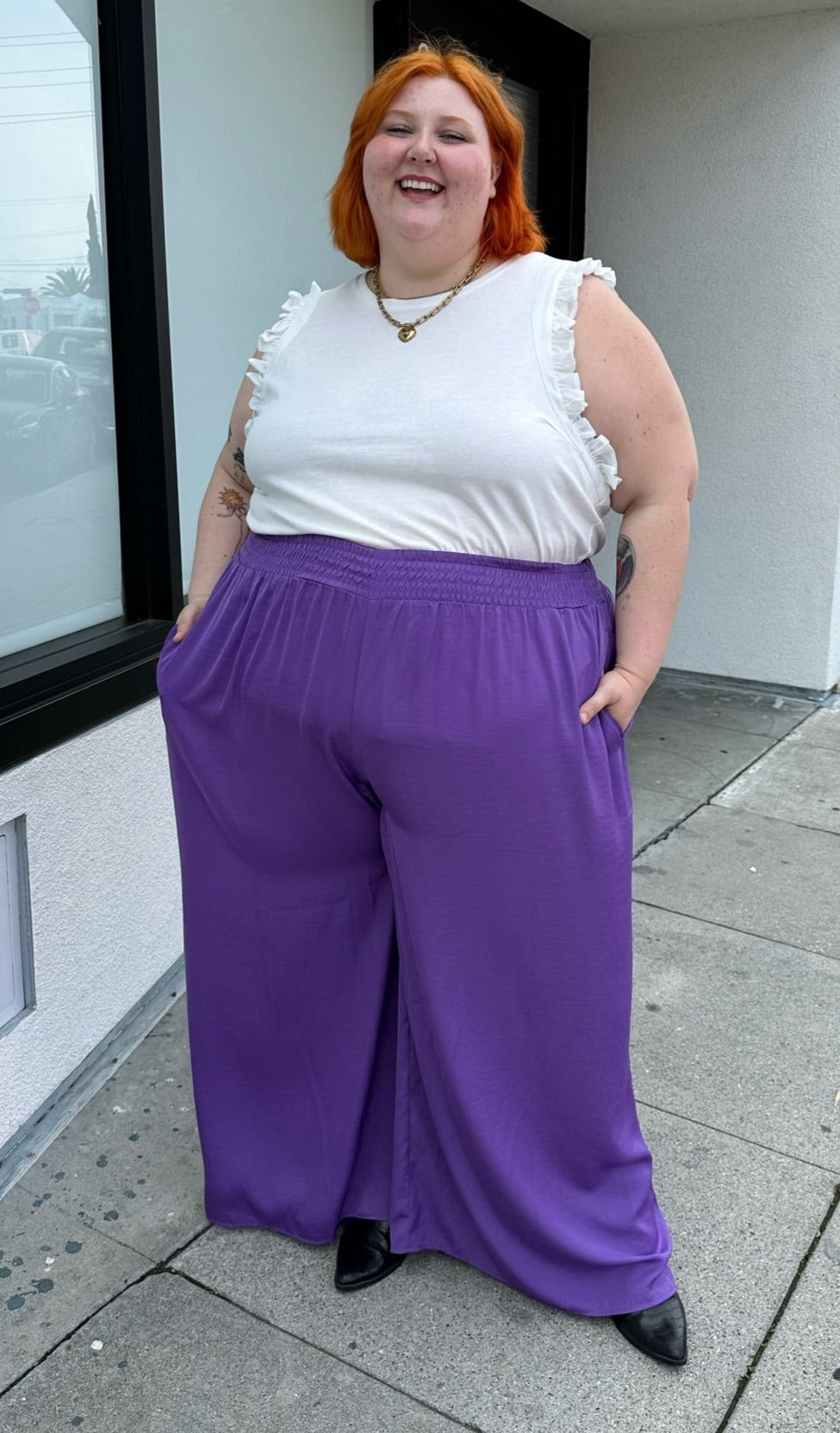 Full-body front view of a pair of size 28 Eloquii purple lightweight wide leg pants with elastic waistband styled with a white ruffled tank and black boots on a size 22/24 model.