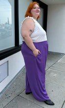 Load image into Gallery viewer, Full-body side view of a pair of size 28 Eloquii purple lightweight wide leg pants with elastic waistband styled with a white ruffled tank and black boots on a size 22/24 model.
