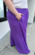 Load image into Gallery viewer, Side view of a pair of size 28 Eloquii purple lightweight wide leg pants with elastic waistband styled with a white ruffled tank and black boots on a size 22/24 model.
