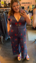 Load image into Gallery viewer, Additional full-body front view of a size L Saint Geraldine navy blue sweater jumpsuit with all-over cherry print, button details, and a tie belt styled with holographic slides on a size 14/16 model.
