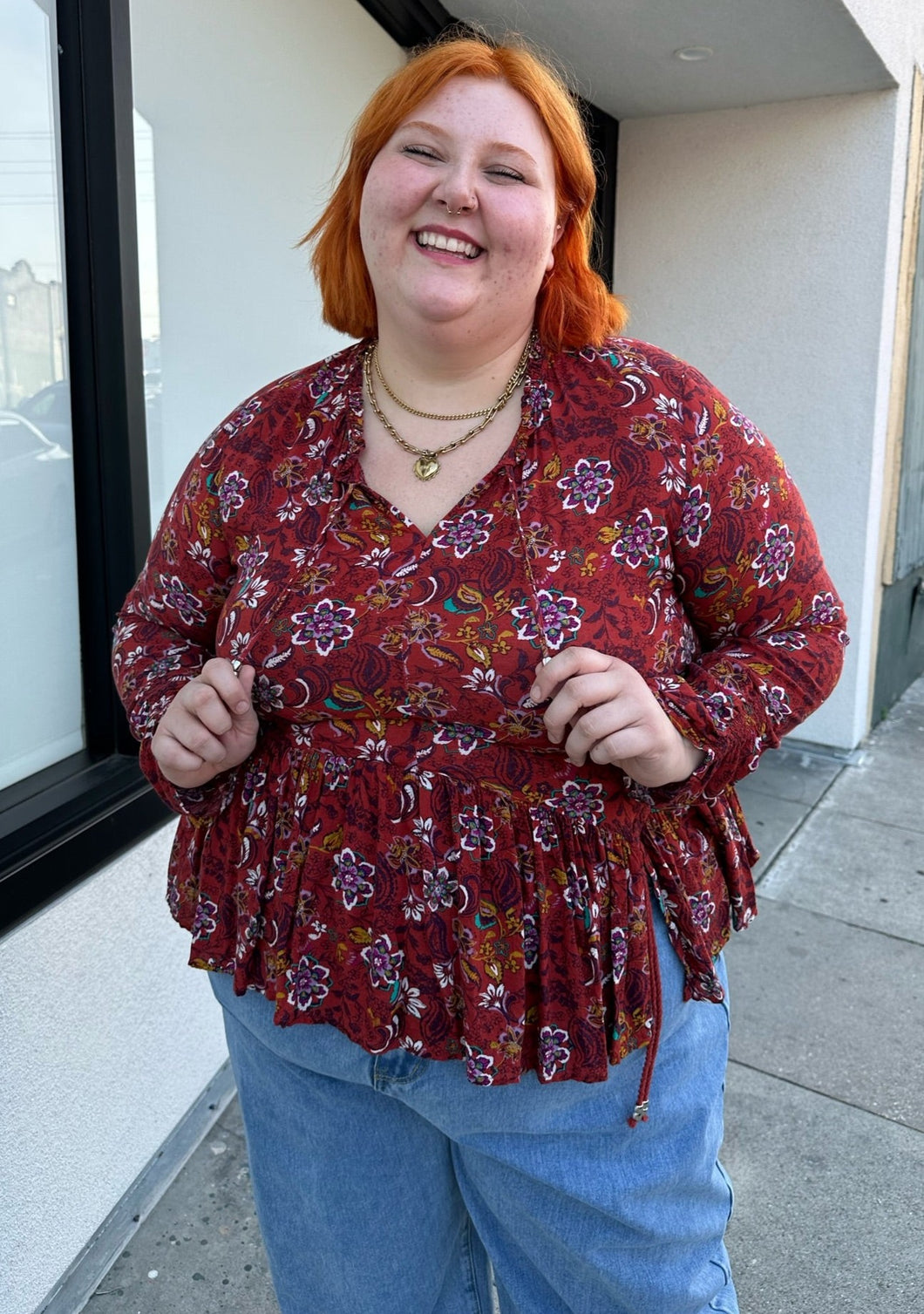 Front view of a size 1/2X deep red, white, teal, yellow, and purple multicolor floral baby doll blouse with long sleeves, a drawstring neckline, and side slits styled with light wash denim on a size 22/24 model.