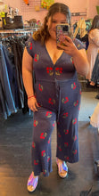 Load image into Gallery viewer, Full-body front view of a size L Saint Geraldine navy blue sweater jumpsuit with all-over cherry print, button details, and a tie belt styled with holographic slides on a size 14/16 model.
