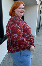 Load image into Gallery viewer, Side view of a size 1/2X deep red, white, teal, yellow, and purple multicolor floral baby doll blouse with long sleeves, a drawstring neckline, and side slits styled with light wash denim on a size 22/24 model.
