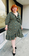 Load image into Gallery viewer, Full-body front view of a size 26 Jason Wu x Eloquii black wrap dress with yellow, blue, green, and orange mini floral pattern and ruffle details styled with black pumps on a size 22/24 model.
