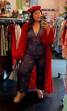 Load image into Gallery viewer, Full-body front view of a size L Saint Geraldine navy blue sweater jumpsuit with all-over cherry print, button details, and a tie belt styled with a red feather-detail duster, a red beret, and brown slides on a size 14/16 model.
