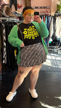 Load image into Gallery viewer, Full-body front view of a size 3X Forever 21 black and white ditsy floral mini circle skirt styled with a black tank, a green cardigan, a black beret, and white crocs on a size 22/24 model.
