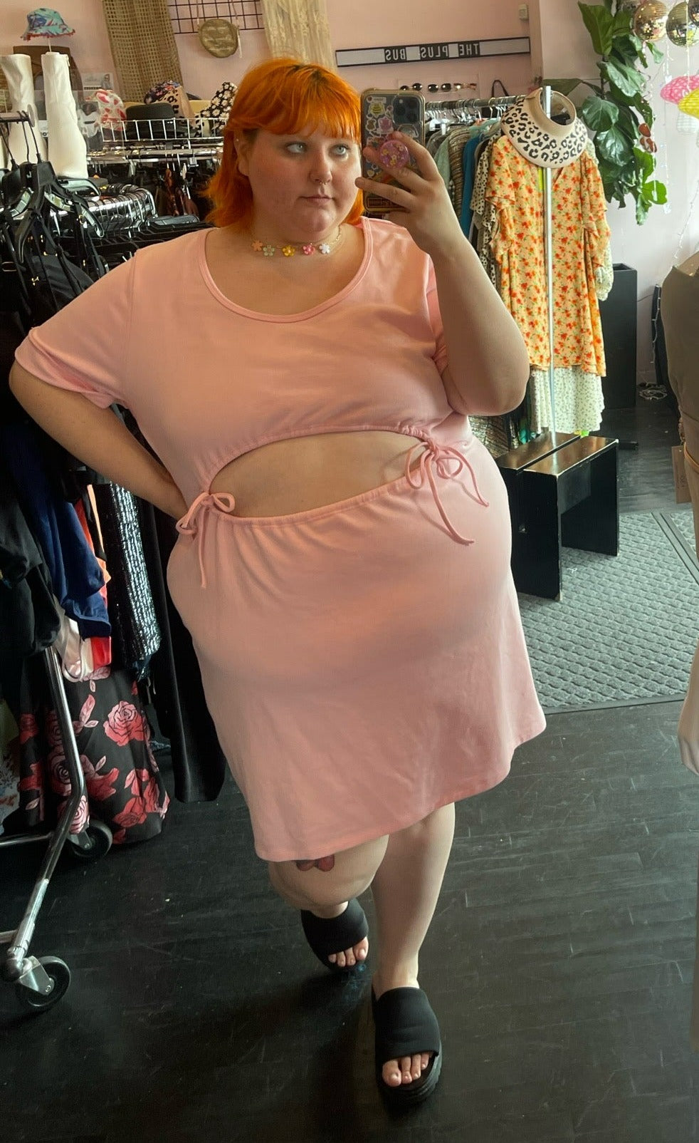 Full-body front view of a size 4 Fashion to Figure baby pink short sleeve sweatshirt-style midi dress with a drawstring belly cut-out styled with a floral choker and black slides on a size 22/24 model.